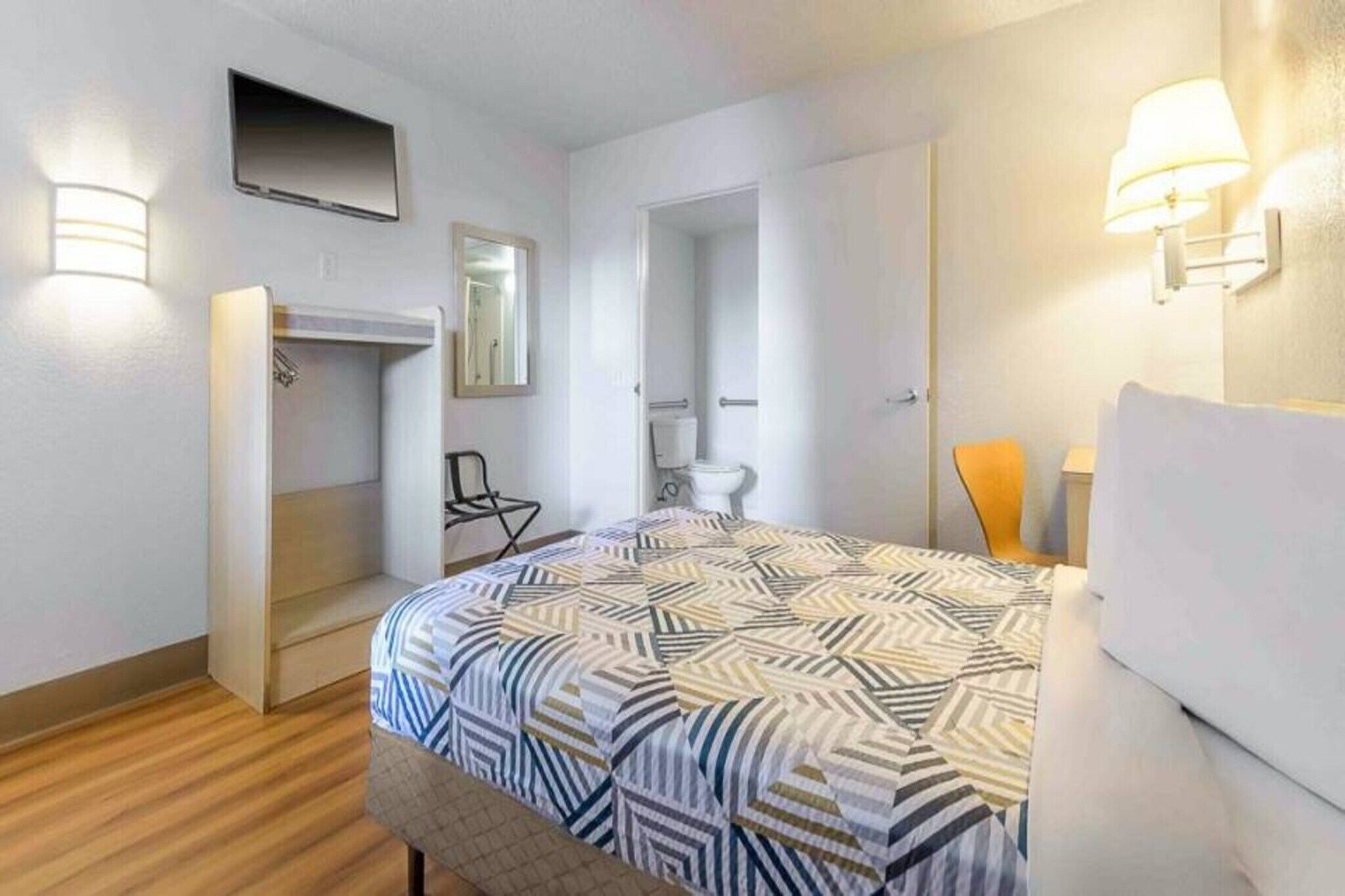 A hotel room with a bed, a chair, a TV, a mirror, and a door leading to a bathroom.