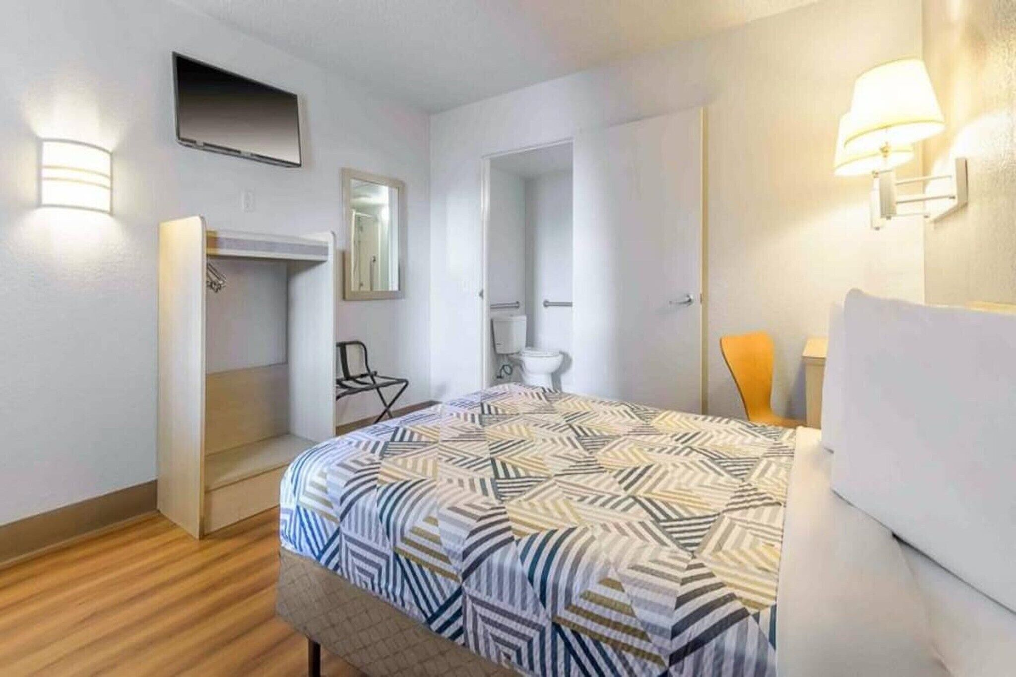 Une chambre d’hôtel équipée d’un lit, d’une chaise, d’une armoire, d’un miroir, de toilettes et d’une télévision.
