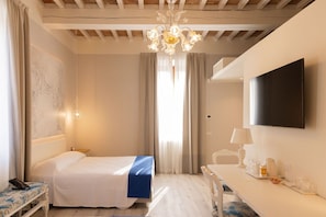 A hotel room with a bed, a desk with a chair, a TV mounted on the wall, and a chandelier.
