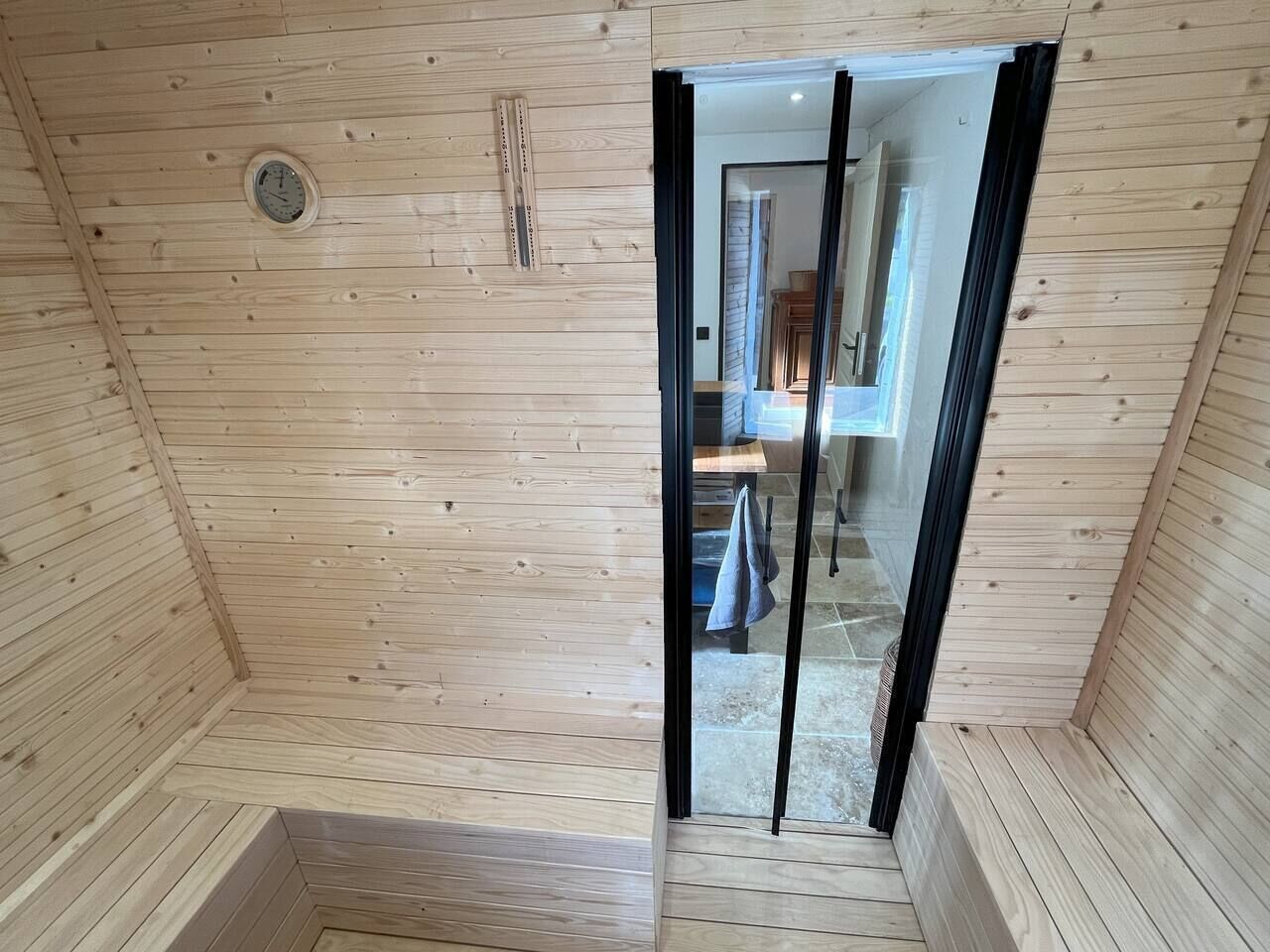 A wooden sauna room with a wooden bench, a clock, and a glass door leading to a bathroom.