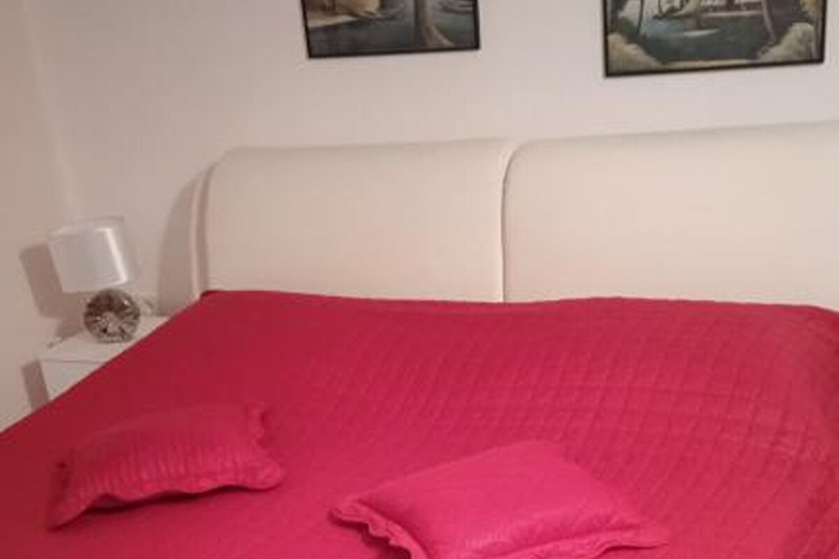 A bed with a red bedspread and pillows, a white headboard, and a nightstand with a lamp and a decorative item.
