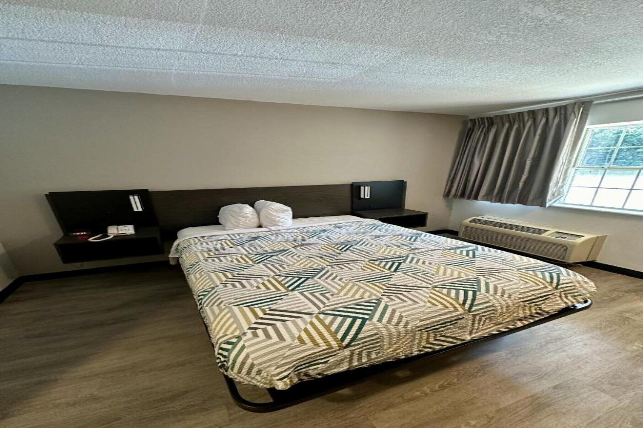 A hotel room with a bed, a nightstand, a heater, and a window with curtains.
