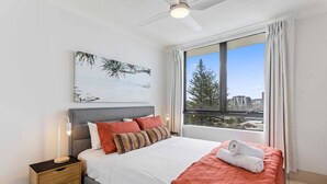 Ein Schlafzimmer mit Bett, Nachttisch, Deckenventilator, Fenster mit Vorhängen und Blick auf die Stadt.