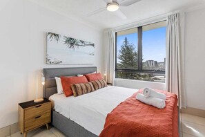 Ein Schlafzimmer mit Bett, Nachttisch, Deckenventilator, Fenster mit Vorhängen und Blick auf die Stadt.