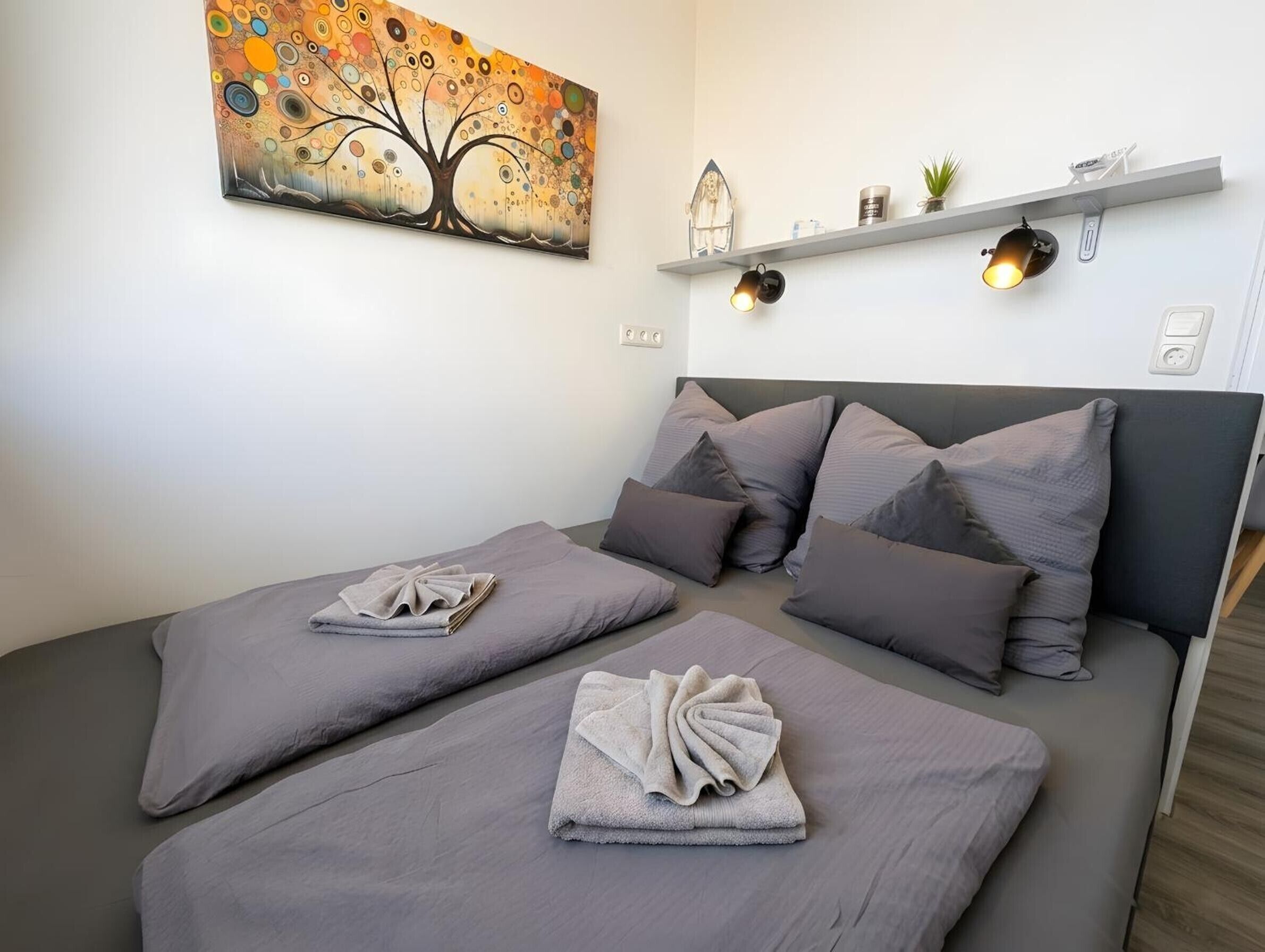 Ein Doppelbett mit grauer Bettwäsche und Kissen, eine Wandablage mit Dekorationselementen und ein Baumgemälde.