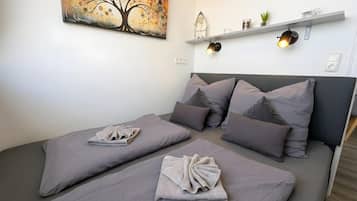 Ein Doppelbett mit grauer Bettwäsche und Kissen, eine Wandablage mit Dekorationselementen und ein Baumgemälde.