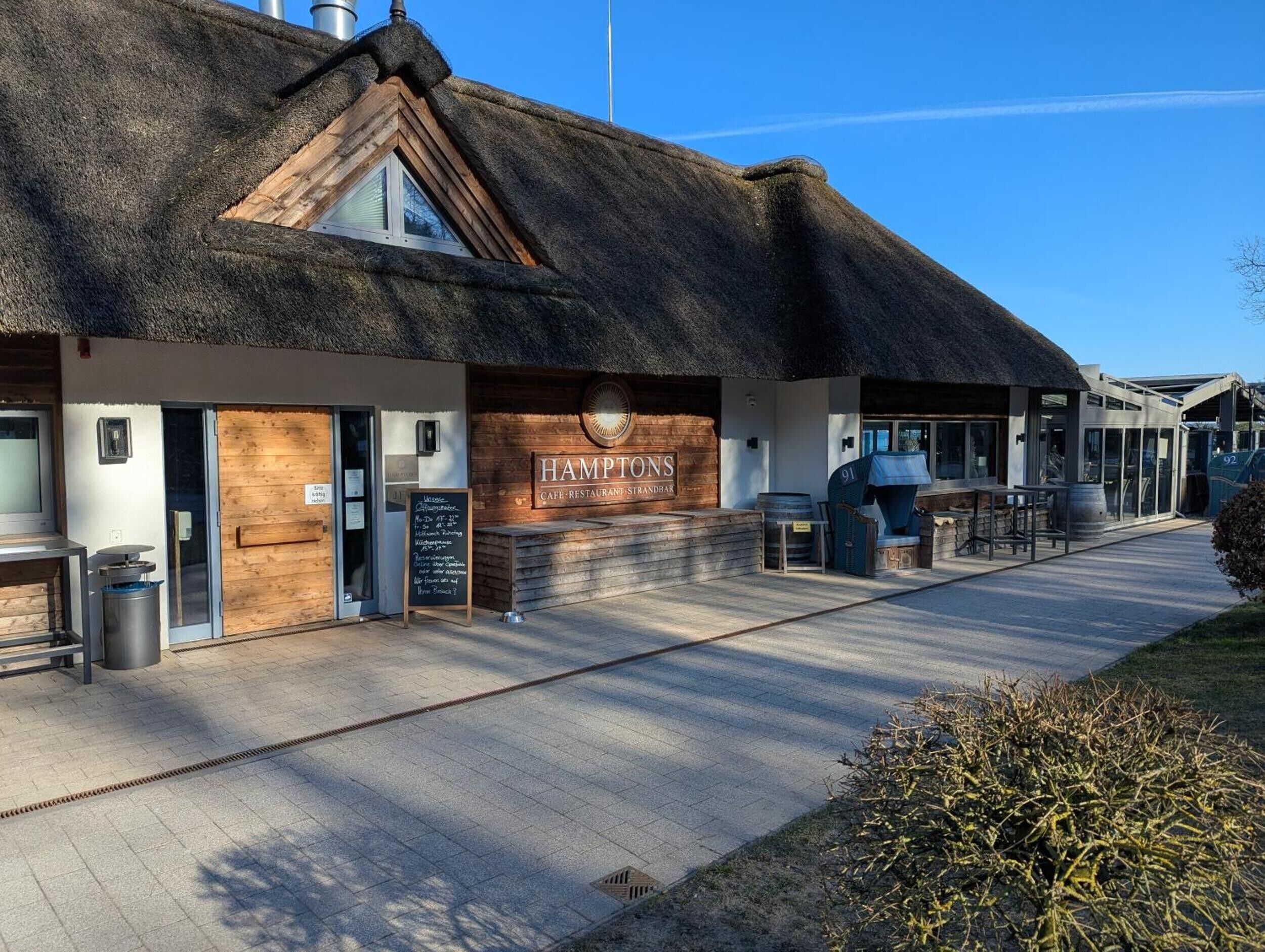 A thatched-roof building with a sign reading 'HAMPTONS CAFE RESTAURANT STRANDBAR'.