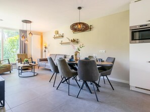 A modern dining area with a round table, grey upholstered chairs, and a hanging light fixture. - 6-person Bungalow With Field Views (Oostkapelle)