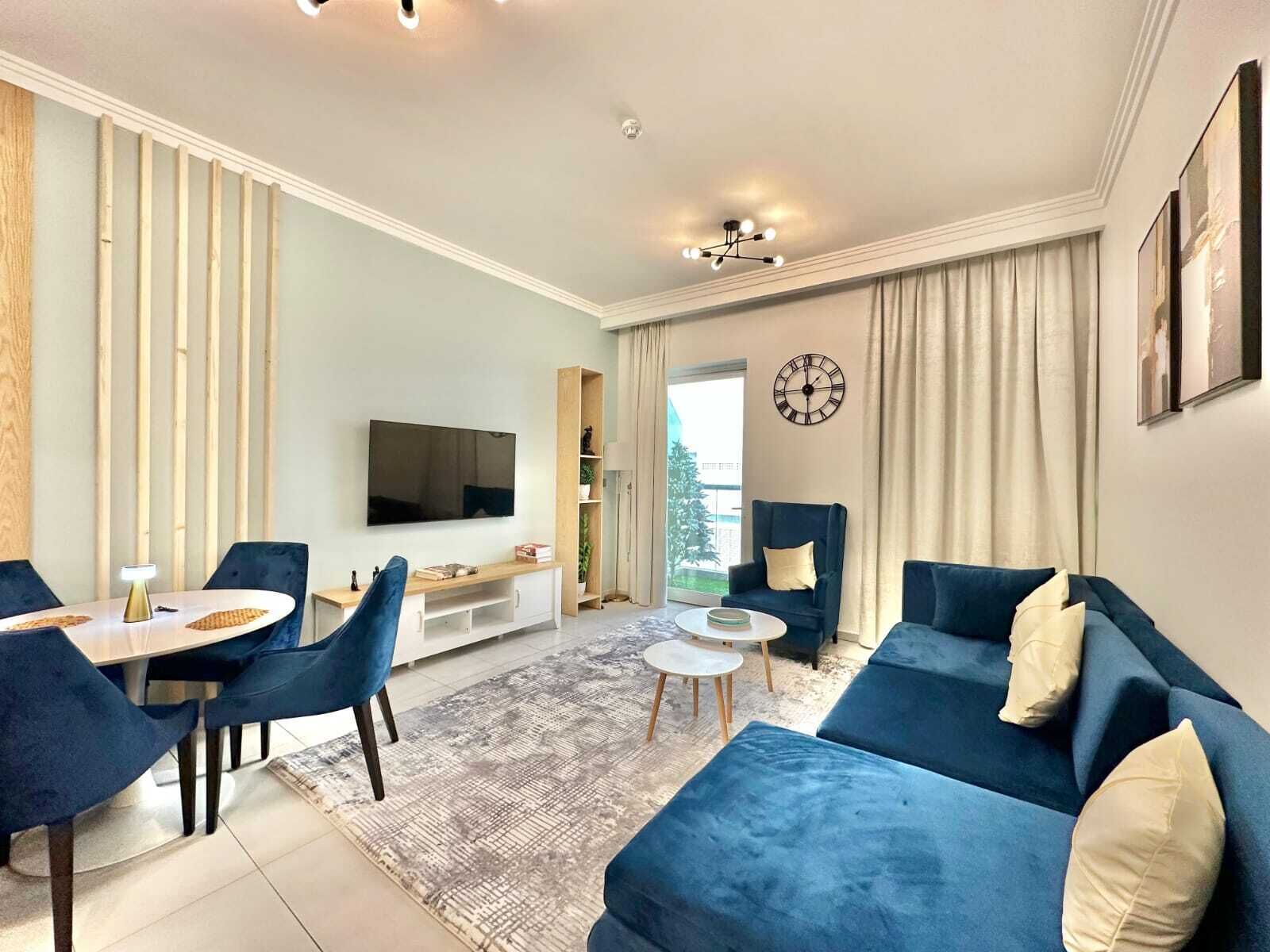 Un salon moderne avec un téléviseur, des canapés bleus, une table à manger avec des chaises et une grande fenêtre.