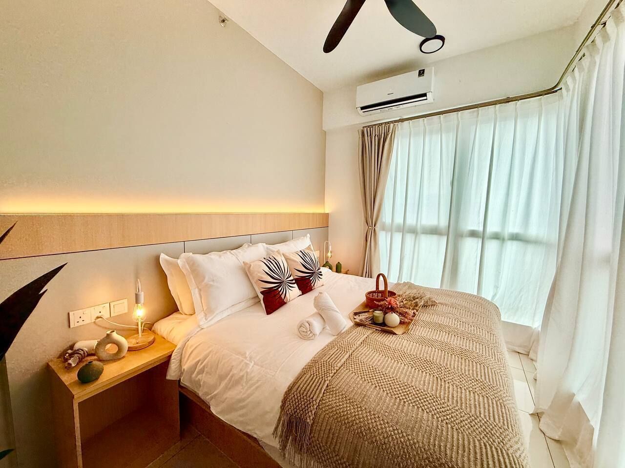 A hotel room with a bed, bedside table, lamp, air conditioning unit, and a window with curtains.