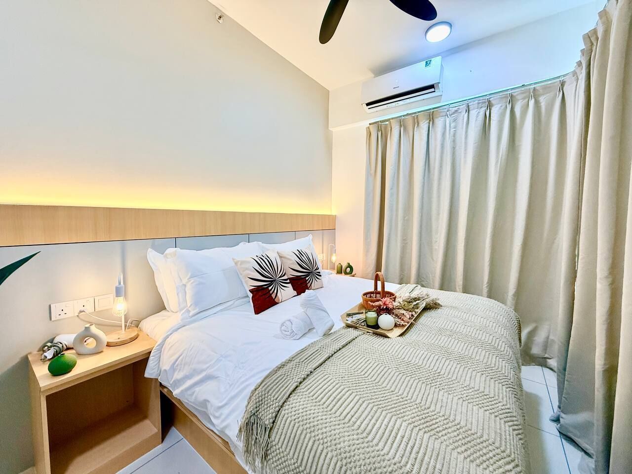 Quarto de hotel com cama, mesa de cabeceira, luminária, aparelho de ar condicionado, cortinas e ventilador de teto.