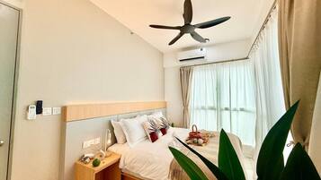 A hotel room with a bed, bedside table, nightstand, ceiling fan, window with curtains, and a potted plant.