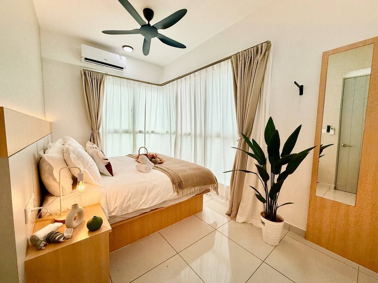 Une chambre moderne comprenant un lit, une table de chevet, une lampe, un climatiseur, un ventilateur de plafond, une grande fenêtre avec des rideaux, une plante en pot et un miroir.