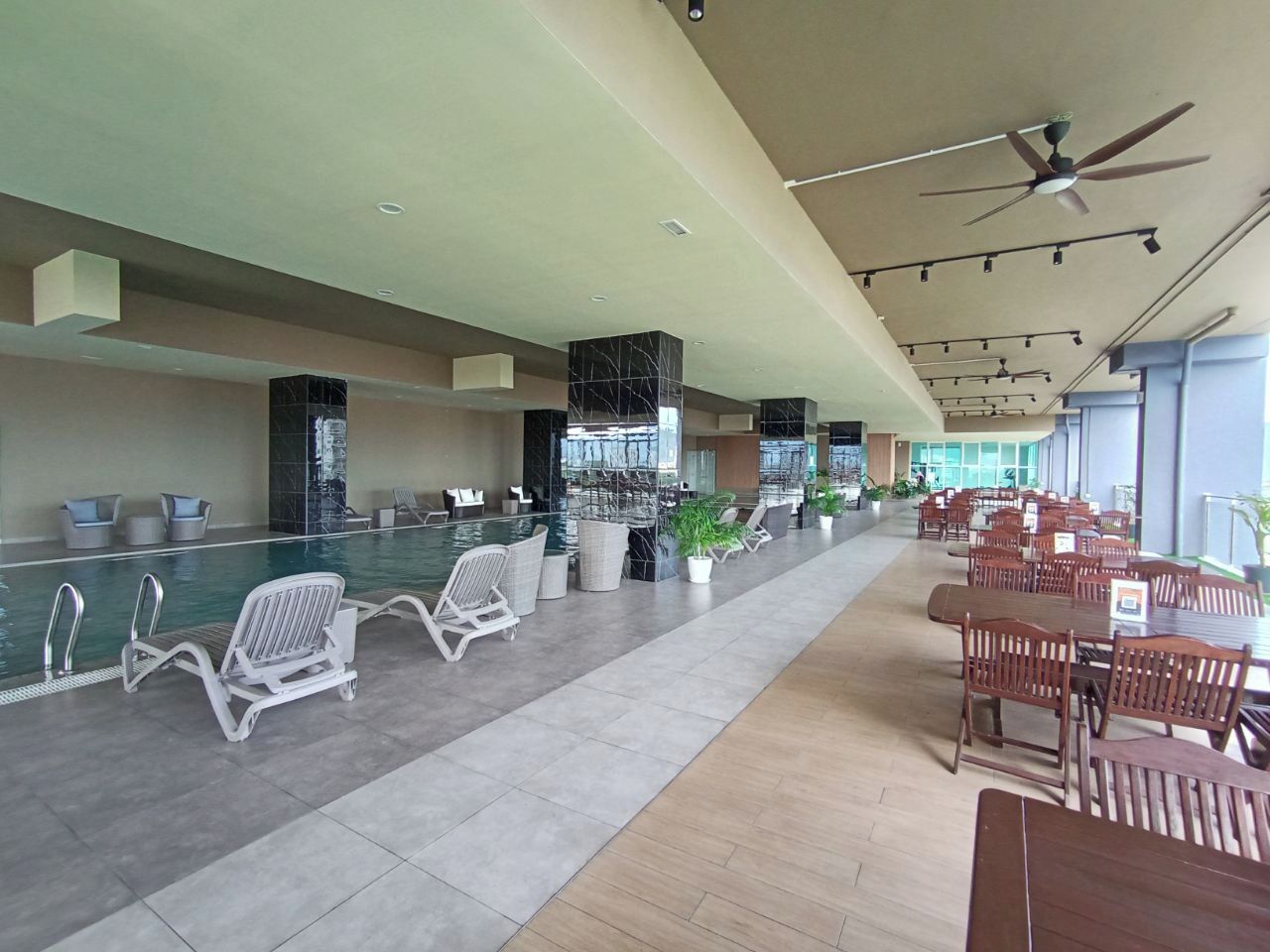 A spacious indoor pool area with lounge chairs and dining tables, a ceiling fan, and large windows.