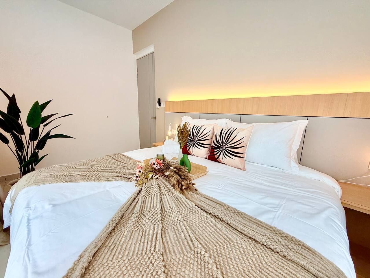 A hotel room with a bed, a nightstand, a potted plant, and a wall-mounted light fixture.