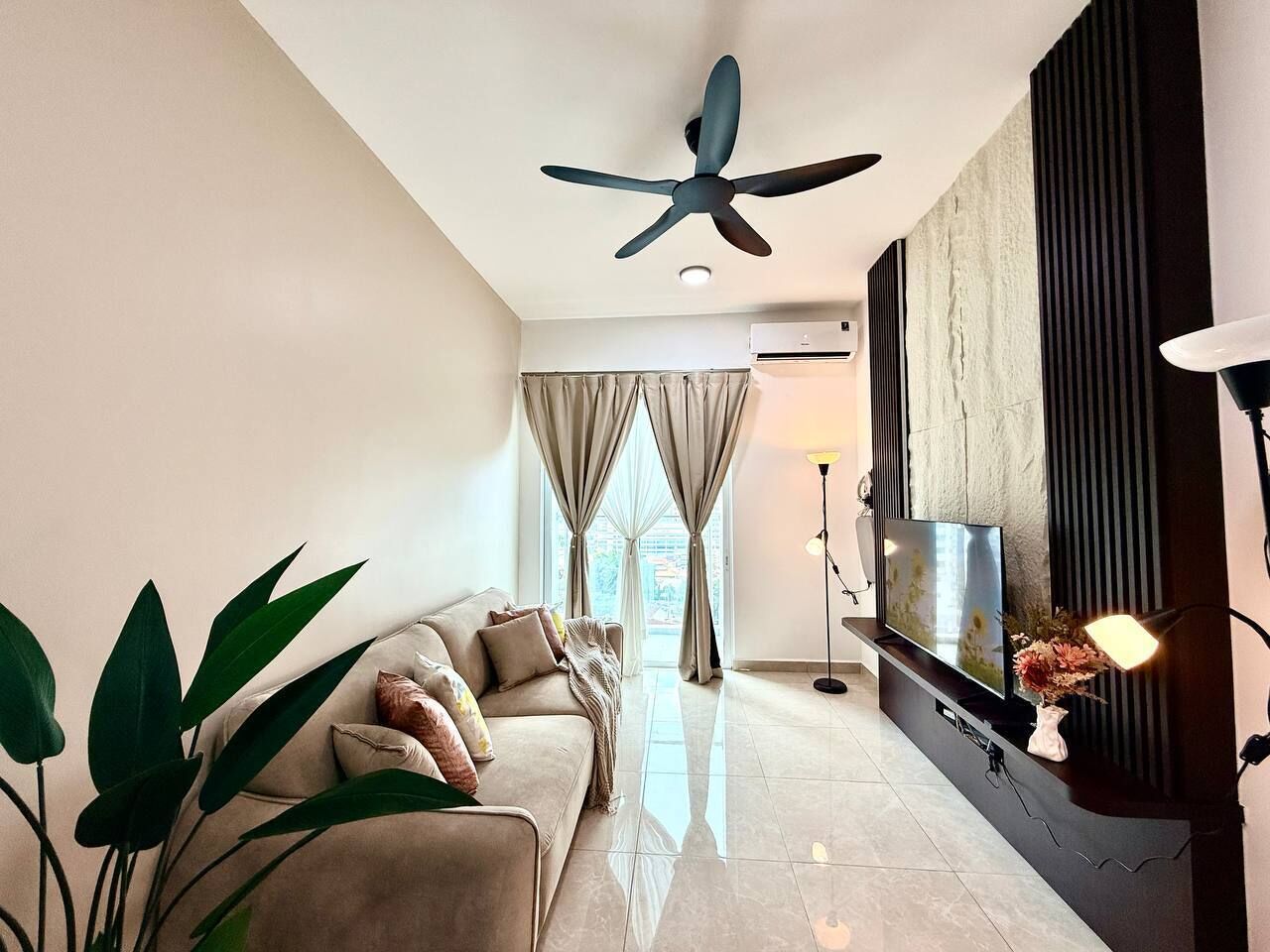 A modern living room with a sofa, TV, ceiling fan, and large windows with curtains.