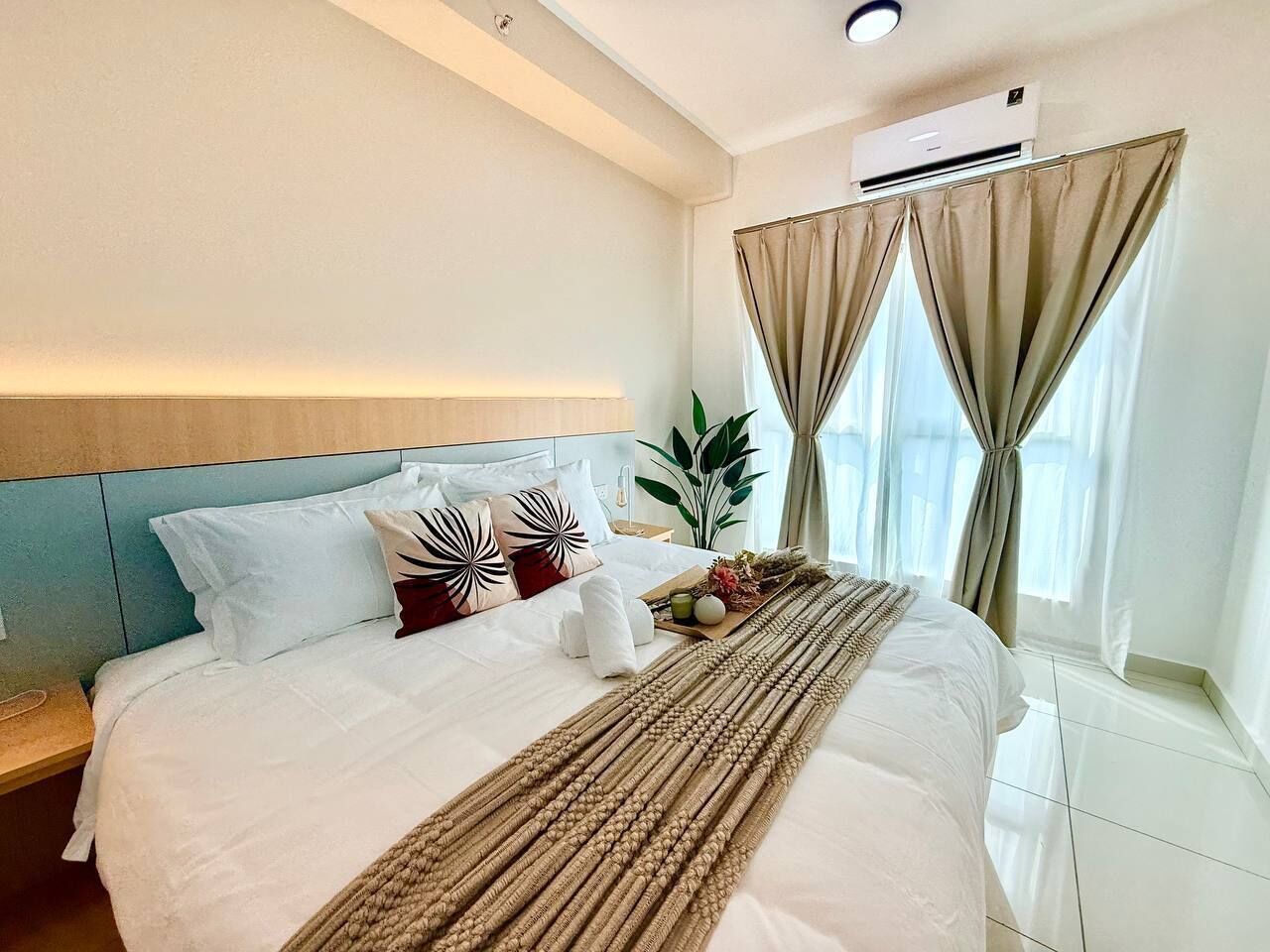 A hotel room with a bed, a headboard, an air conditioning unit, a window with curtains, and a bedside table.
