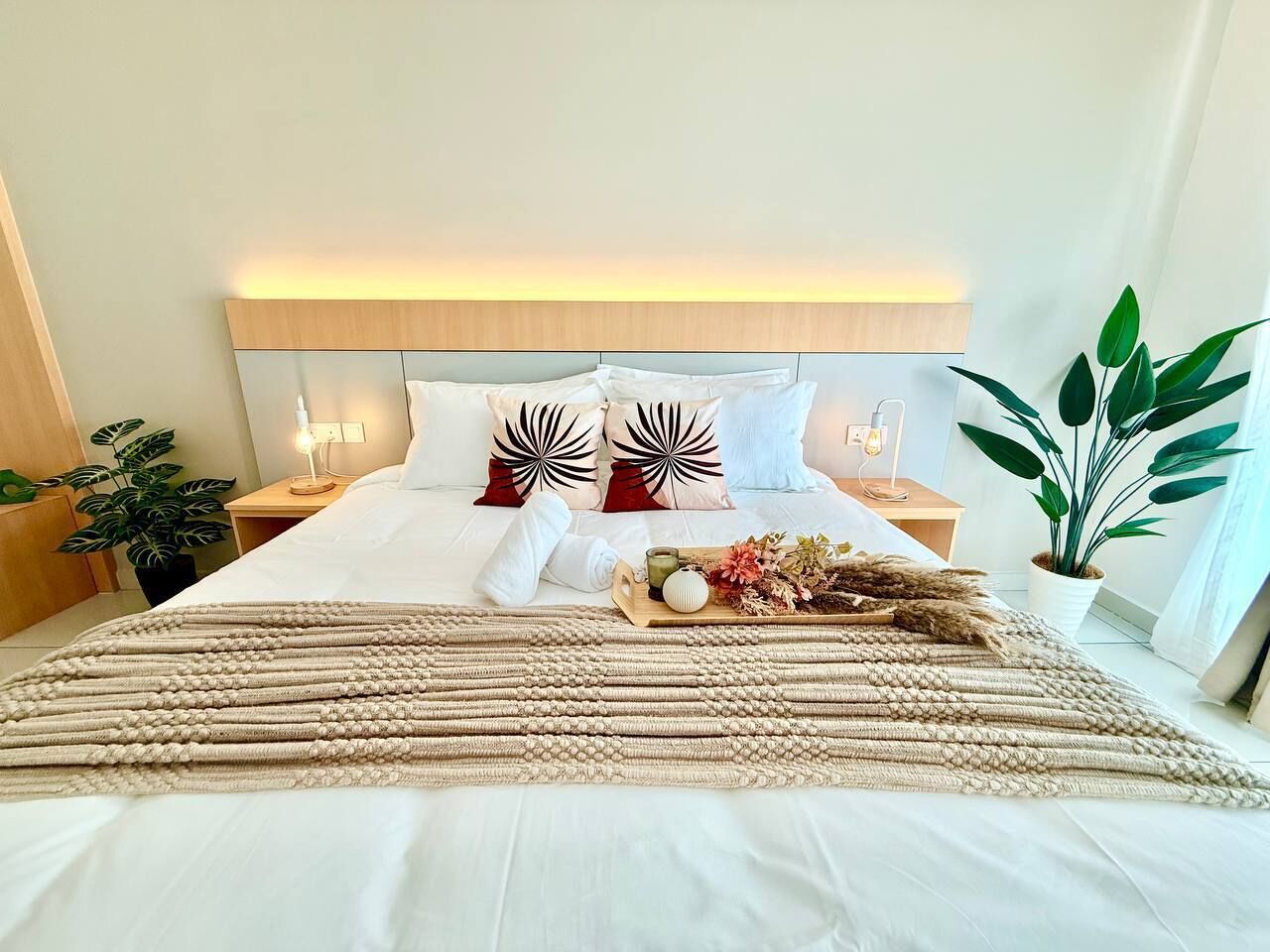 A neatly made bed with a headboard, a bedside table with a lamp, a potted plant, and a tray with decorative items.