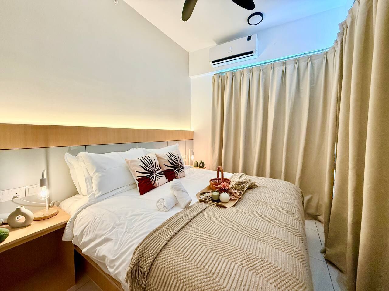 A hotel room with a bed, bedside table, lamp, air conditioning unit, curtains, and a tray with snacks.