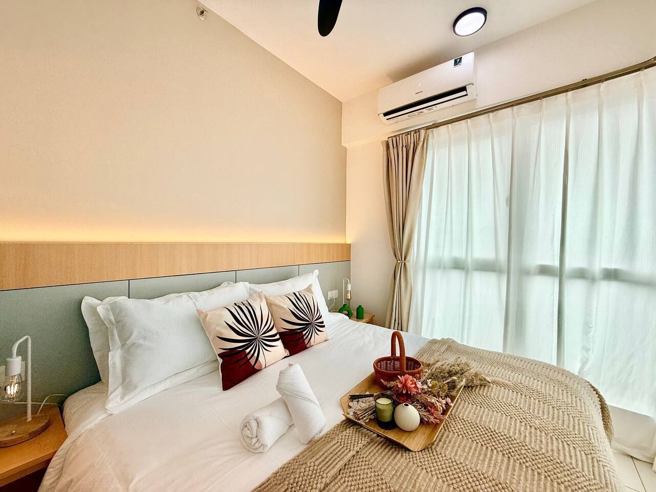 A hotel room with a bed, an air conditioning unit, a nightstand, a lamp, and a window with curtains.
