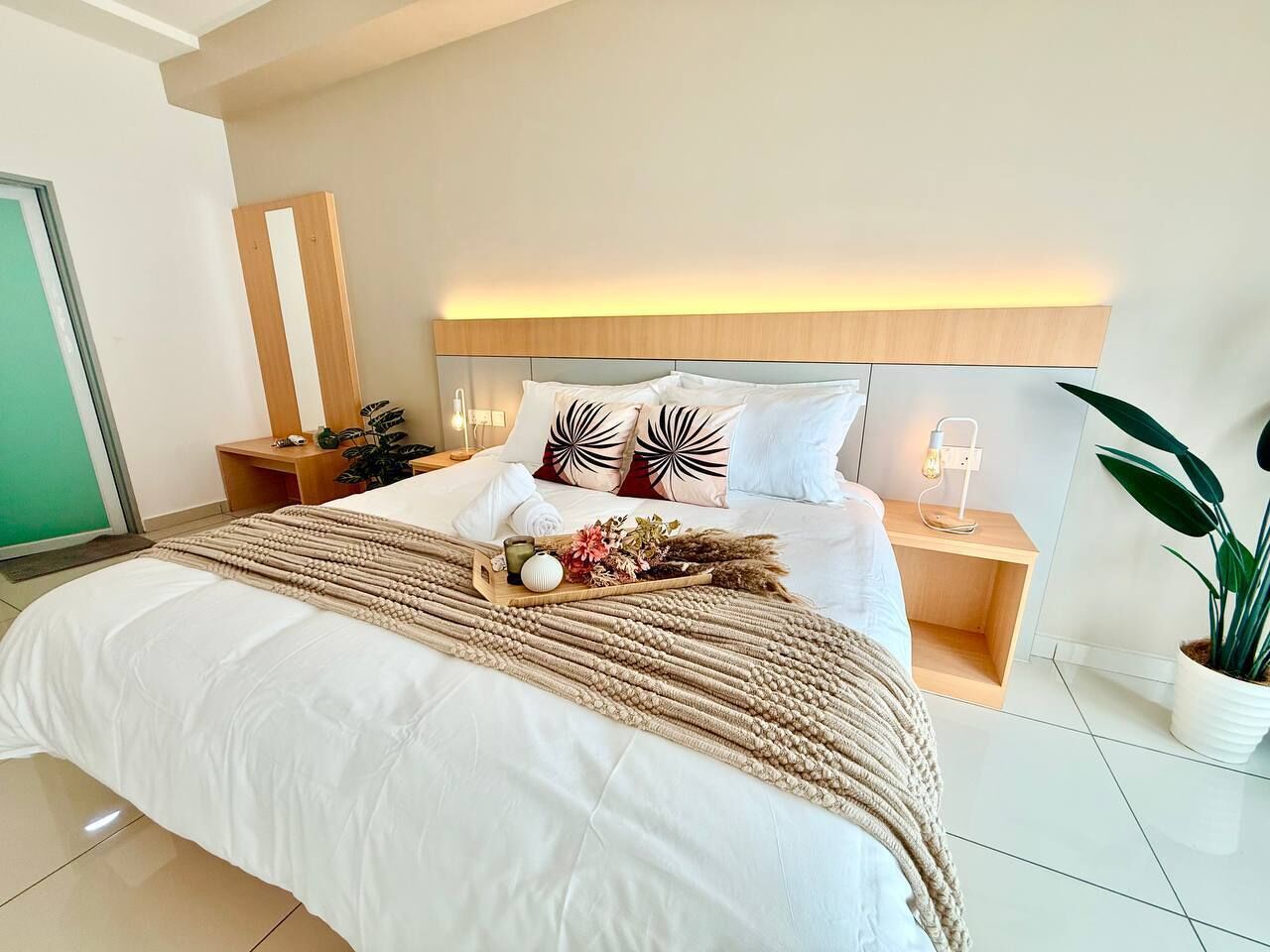 A modern bedroom with a large bed, bedside tables, a nightstand, a wall-mounted light, and a potted plant.