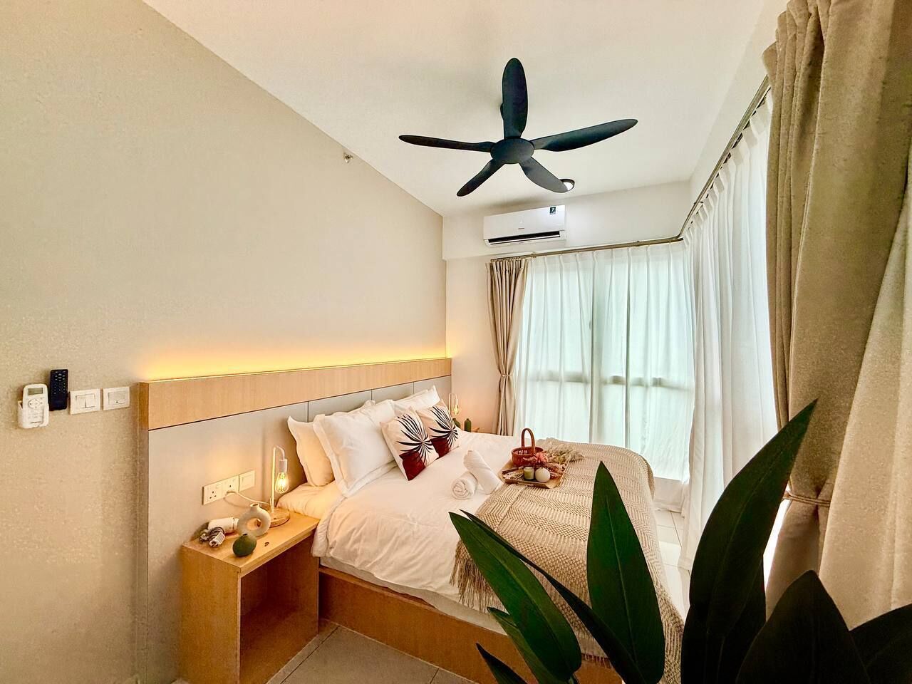 Une chambre d’hôtel comprenant un lit, une table de chevet, une commode, un ventilateur de plafond, un appareil de climatisation et une fenêtre avec des rideaux.