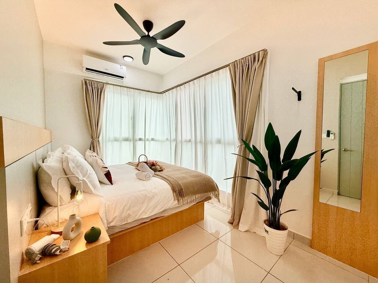 A modern hotel room with a bed, bedside table, air conditioning unit, ceiling fan, large window with curtains, potted plant, and a mirror.