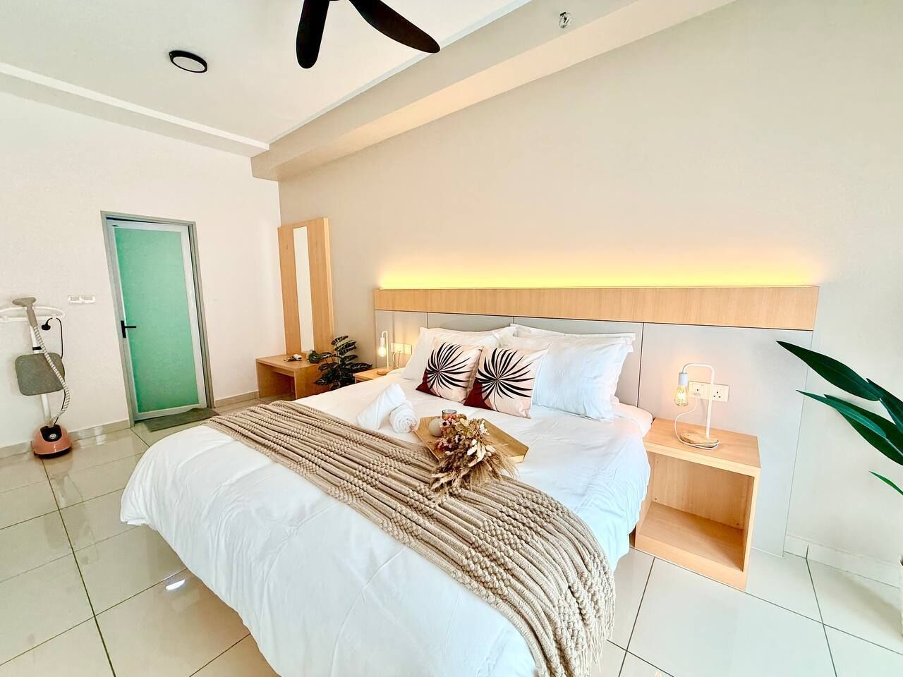 Una habitación de hotel moderna con una cama grande, mesitas de noche, una lámpara de noche, una puerta verde y un ventilador de techo.