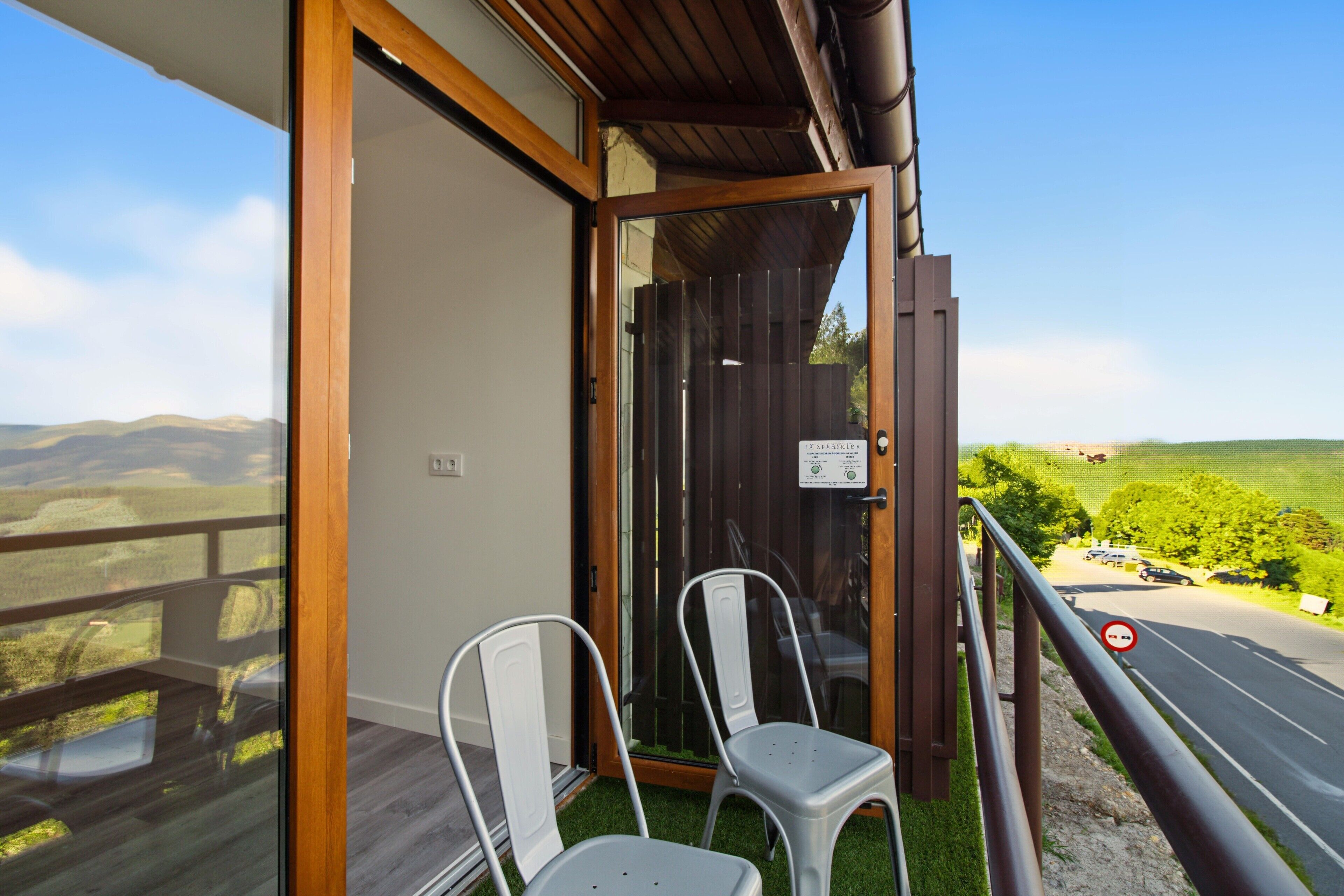 A balcony with two chairs, a wooden door, and a view of a road and greenery.