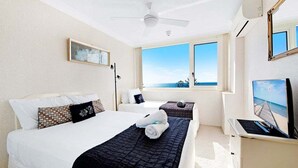 Ein Hotelzimmer mit zwei Betten, einem Deckenventilator, einer Klimaanlage, einem Fernseher und einem Fenster mit Blick aufs Meer.