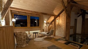Um quarto de sótão acolhedor com parede de pedra, vigas de madeira, escada de vidro e um espaço de estar com mesa e cadeiras.