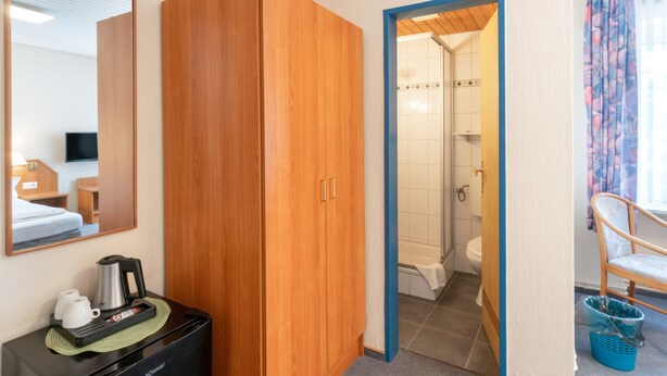 Habitació d'hotel amb un armari de fusta, una màquina de cafè, un mirall, una zona de dutxa i una cadira.