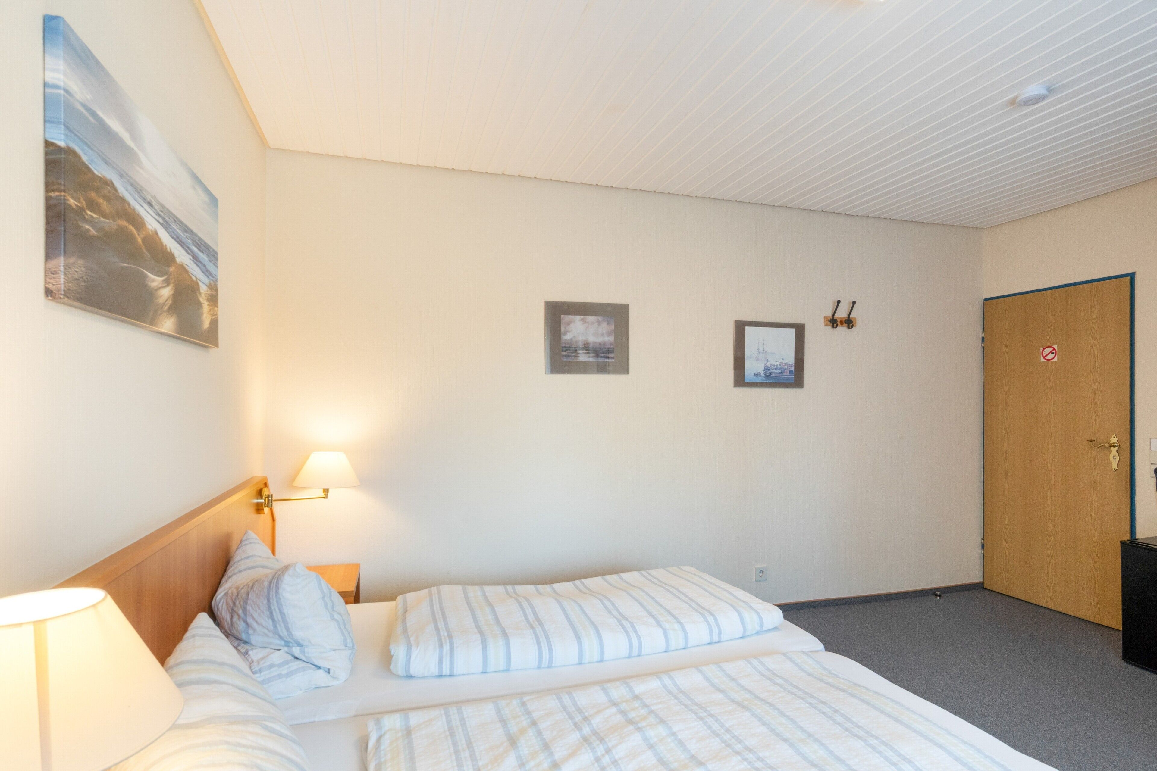 A hotel room with a bed, a nightstand, a lamp, two framed pictures on the wall, and a door with a 'no entry' sign.