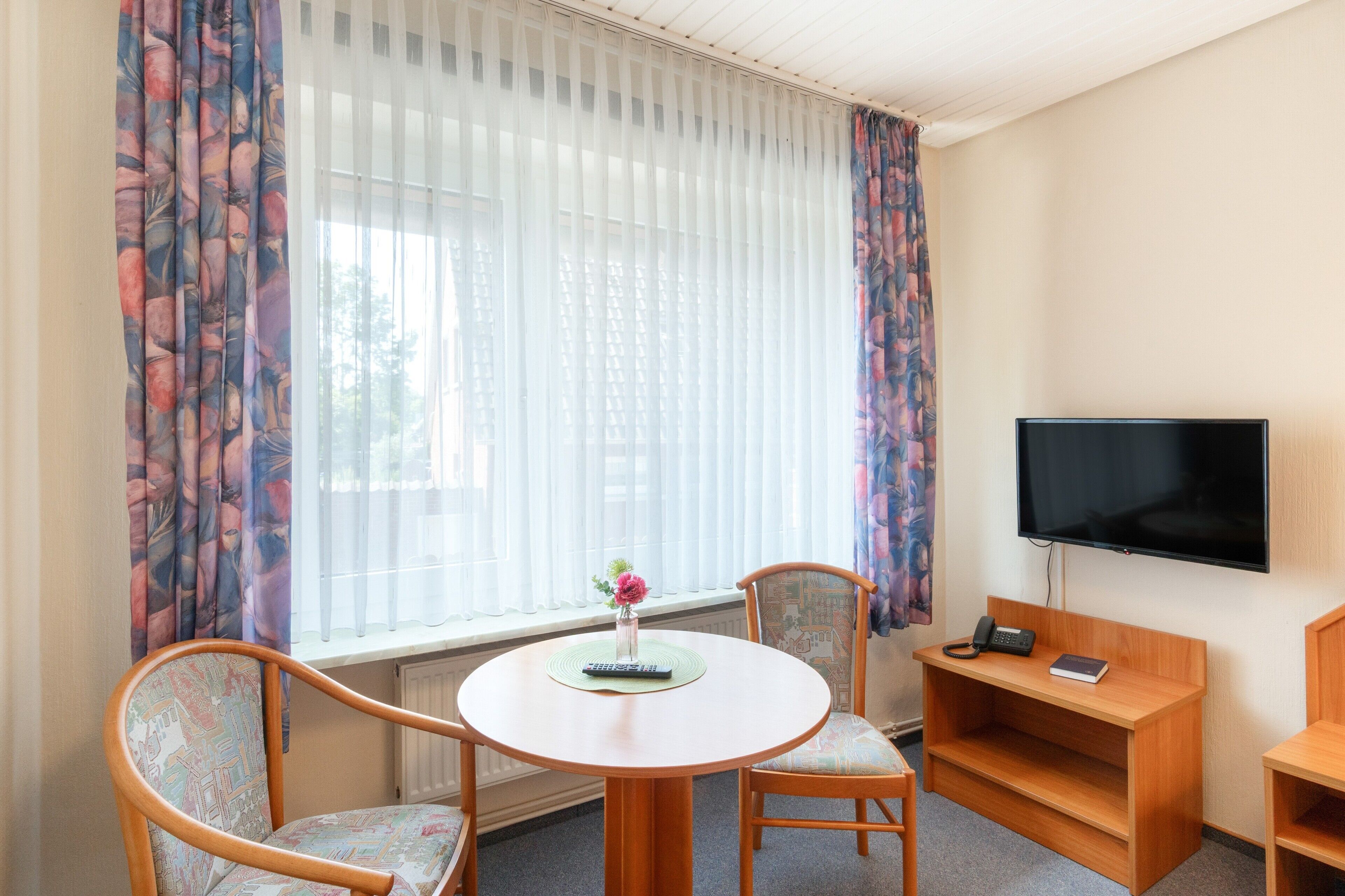 Ein Hotelzimmer mit rundem Tisch, zwei Stühlen, einem Fernseher, einem Telefon und einem Fenster mit leichtem Vorhang.