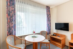 Ein Hotelzimmer mit rundem Tisch, zwei Stühlen, einem Fernseher, einem Telefon und einem Fenster mit leichtem Vorhang.