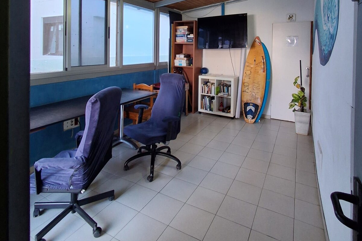 A room with a wooden ceiling, a desk, two chairs, a bookshelf, a surfboard, and a wall-mounted TV.