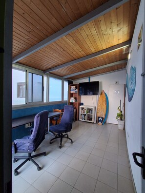 A room with a wooden ceiling, a desk, two chairs, a bookshelf, a surfboard, and a wall-mounted TV. - Pura Vida by Nest Hostel (Las Palmas de Gran Canaria)