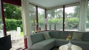 A living room with a sectional sofa, a television on a stand, a small table with a vase, and a view of greenery outside. - Quiet Coastal Bungalow in Oostkapelle (Oostkapelle)