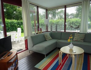 Ett vardagsrum med en hörnsoffa, en tv på ett tv-bord, ett litet bord med en vas och utsikt över grönskan utanför.