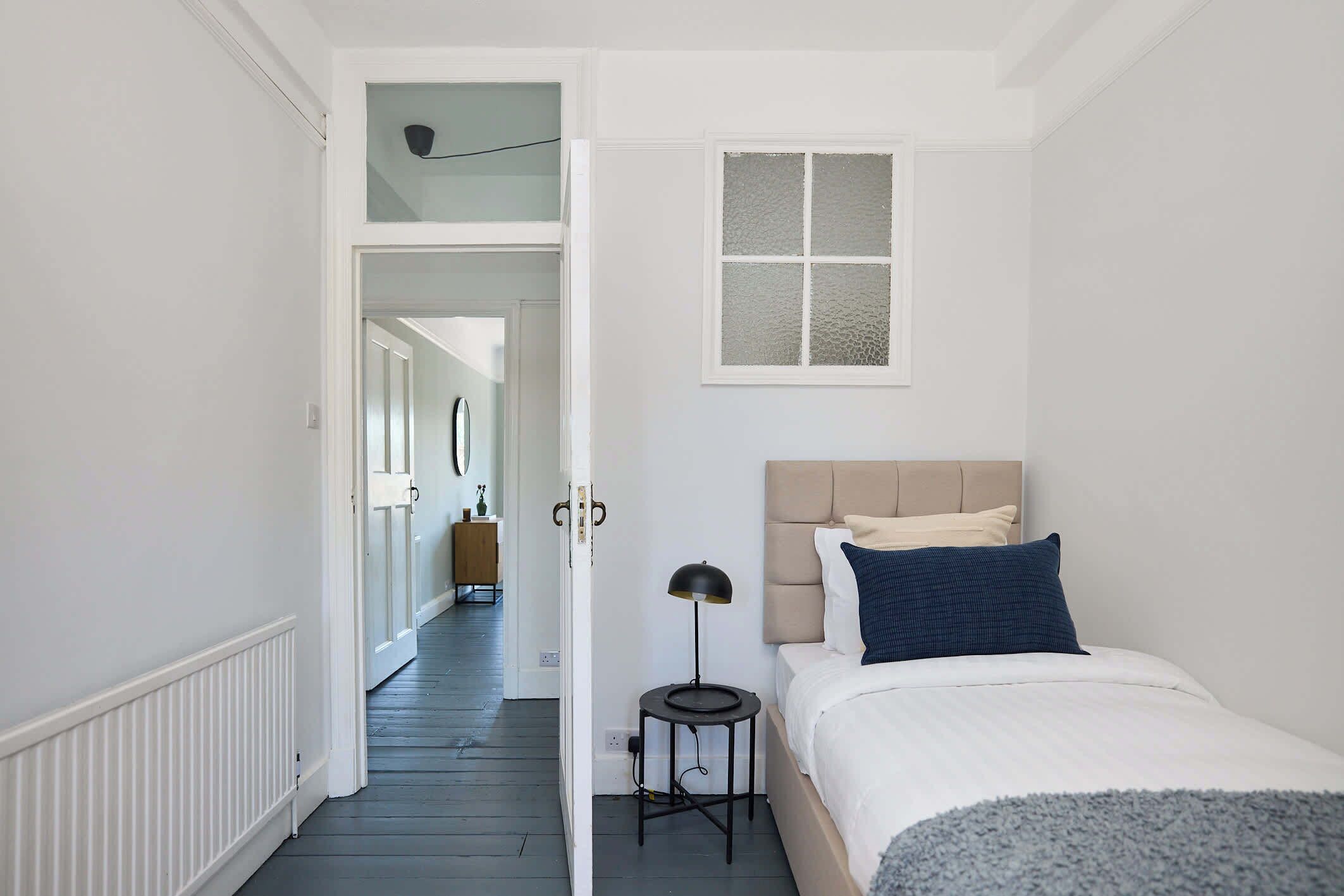 A bedroom with a bed, a window, a lamp, and a door leading to another room.