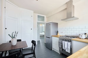 Eine kleine Küche mit Herd, Backofen und Kühlschrank. Ein Esstisch mit Stühlen und einem Blumentopf sind ebenfalls vorhanden.