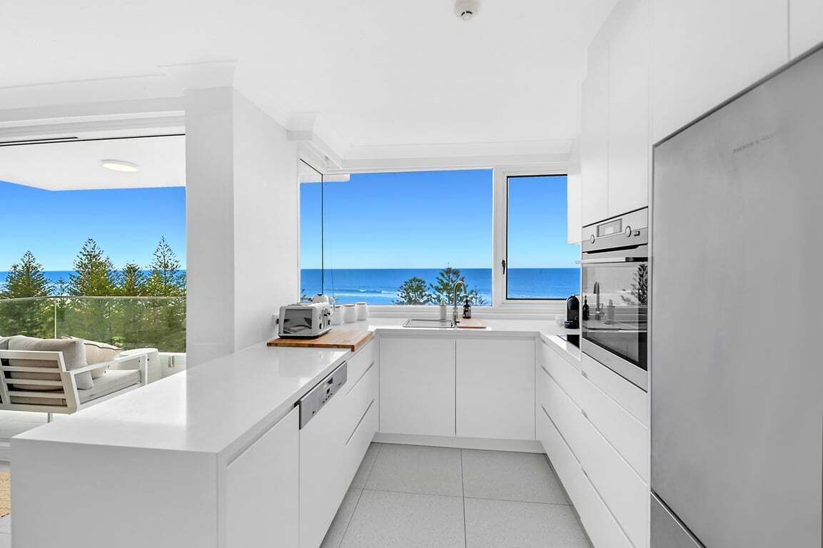 Uma cozinha moderna com armários brancos, forno embutido e uma grande janela com vista para o mar.