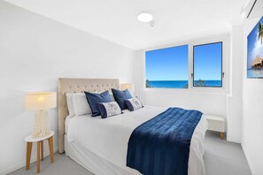 Quarto com cama, mesa de cabeceira, luminária, janela com vista para o mar e um quadro na parede.