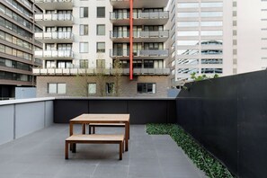 Un'area patio esterna con tavolo e panche in legno, circondata da un muretto basso e un edificio alto sullo sfondo.