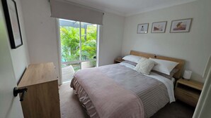 Een slaapkamer met een bed, nachtkastje, stoel, raam met uitzicht op groen, en een deur.