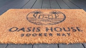 Een deurmat met de tekst 'OASIS HOUSE', een houten deurkozijn en een donkere vloer.