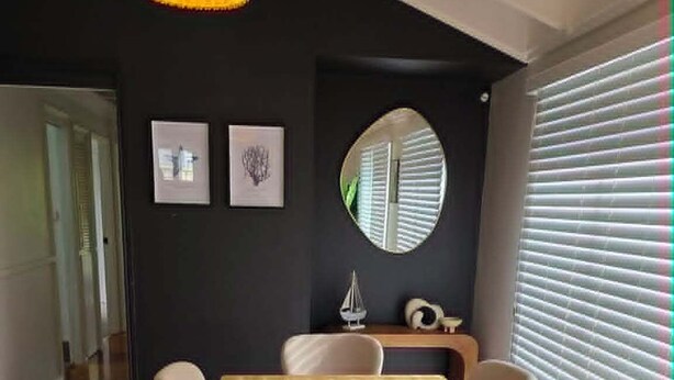 Een eethoek met een houten tafel, zes stoelen, een geweven hanglamp, ingelijste kunstwerken, een ronde spiegel en een raam met jaloezieën.