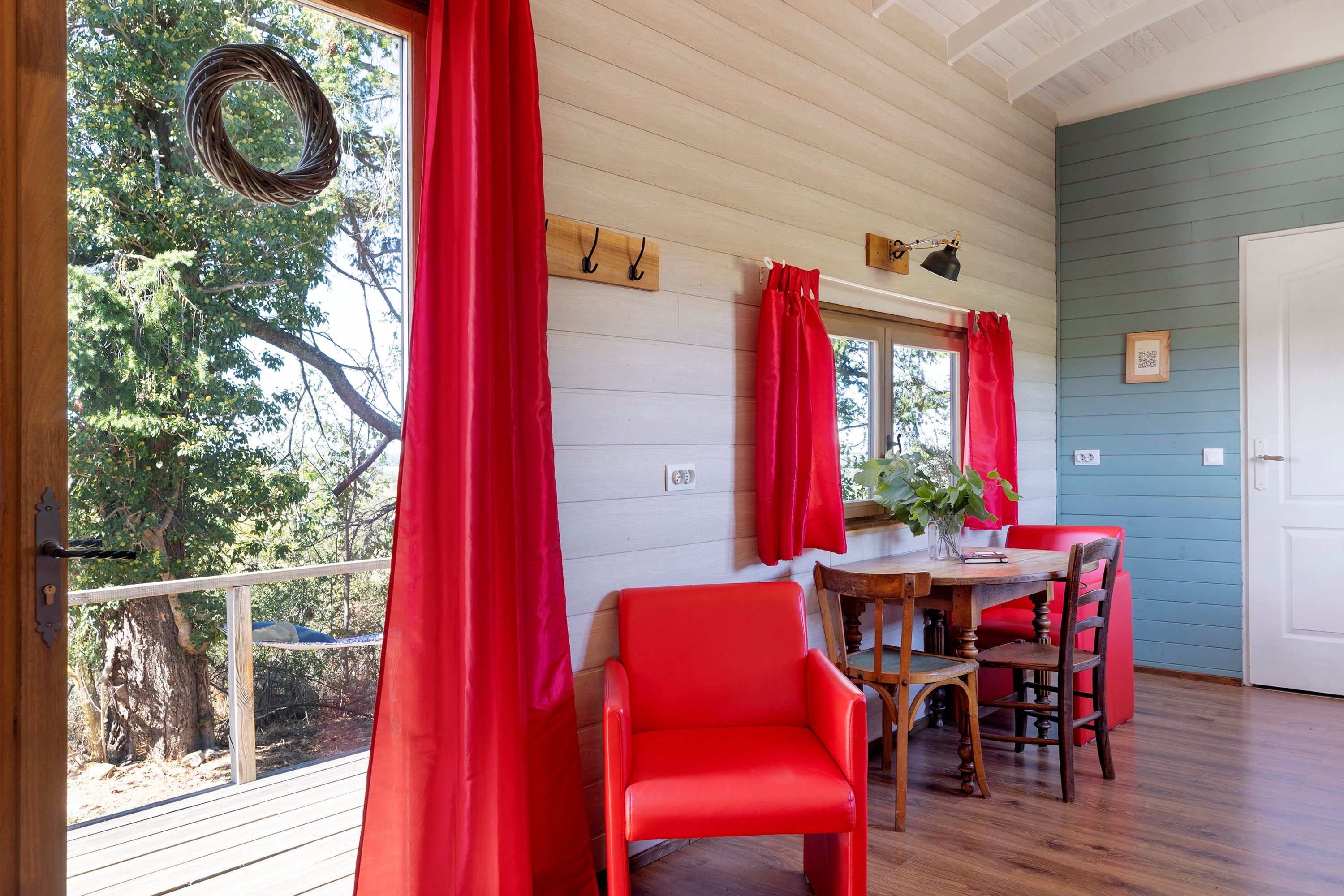 A room with a wooden deck, red curtains, a dining table with chairs, and a view of trees.