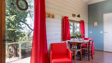 A room with a wooden deck, red curtains, a dining table with chairs, and a view of trees.