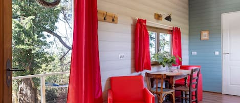 Una habitación con terraza de madera, cortinas rojas, una mesa de comedor con sillas y vista a árboles.